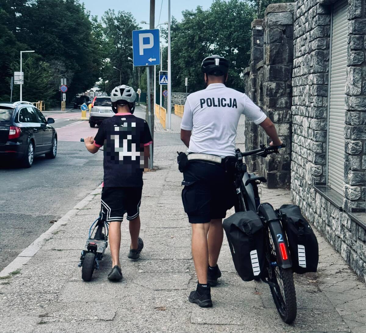 Policjant prowdzący rower, dziecko prowadzące hulajnogę 