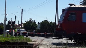 czerwony  pociąg przejeżdża przez torowisko, zapory przejazdu  są opuszczone, przed zaporami stoją samochody, policjant i dwie kobiety  w pomarańczowych kamizelkach odblaskowych.