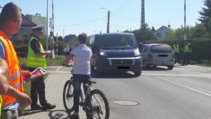 przed torowiskiem stoi mężczyzna w pomarańczowej  kamizelce, policjant i  chłopiec na rowerze. przez torowisko  przejeżdżają samochody