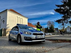 Na zdjęciu znajduje się radiowóz Policji a obok niego stoi umundurowana policjantka z ruchu drogowego. Radiowóz jest na parkingu.