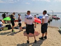 Na plaży dwóch policjantów stoi tyłem obok nich widać dzieci w kamizelkach ratunkowych